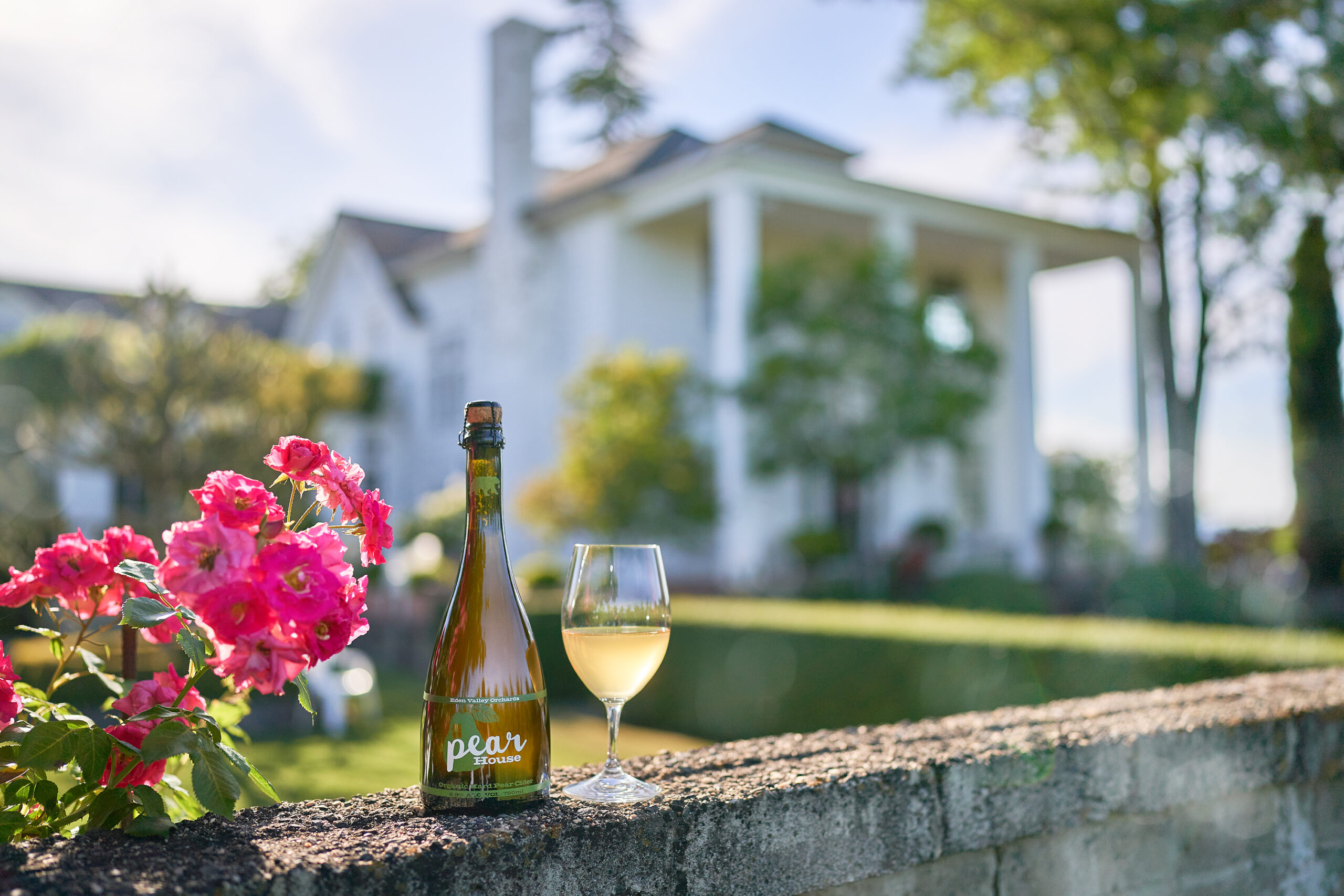 cider bottle, glass of cider and pink flowers in a vase sit on edge of wall in front of historic mansion
